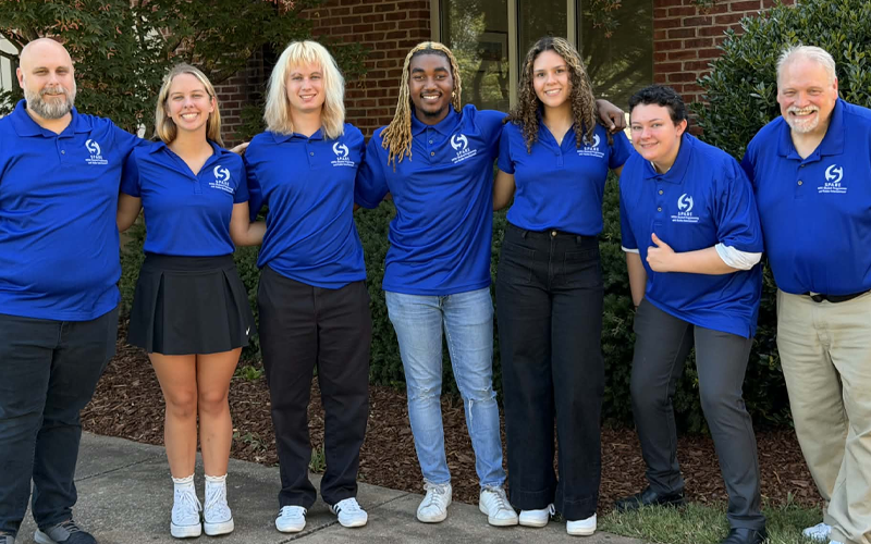 Seven people standing together as a group outside and smiling while wearing blue polos